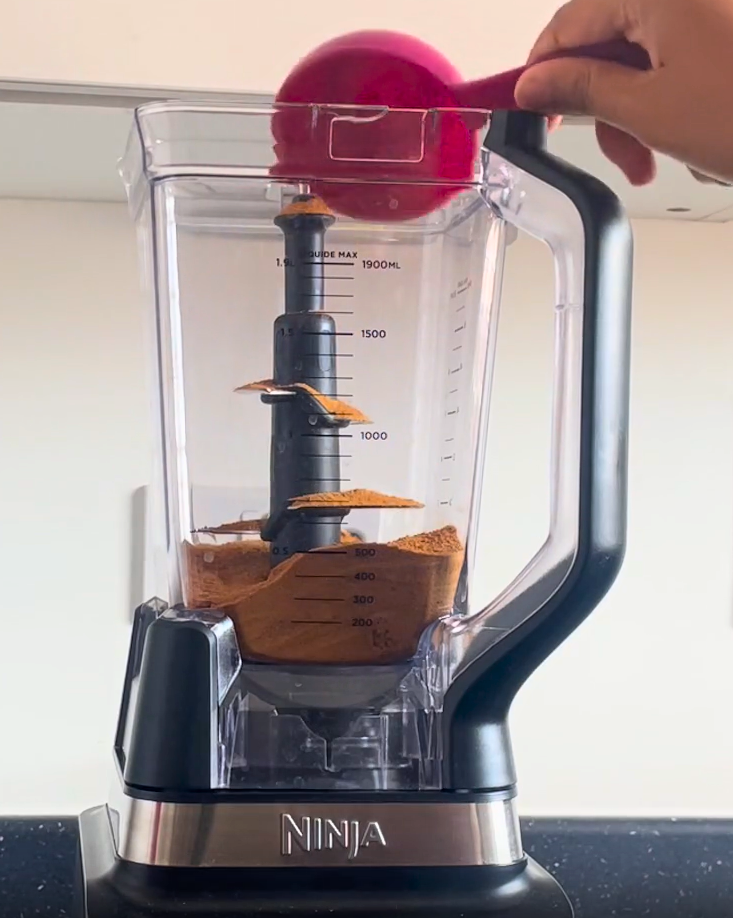 Hand holding pink scoop adding coconut sugar into Ninja blender