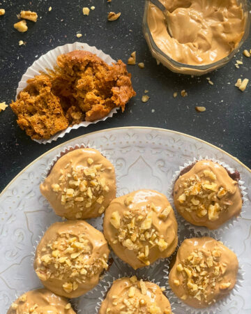 Torn carrot cake muffin and iced carrot cake muffins on decorative plate and a bowl of coconut sugar cream cheese icing with a spoon and chopped walnuts on a black counter