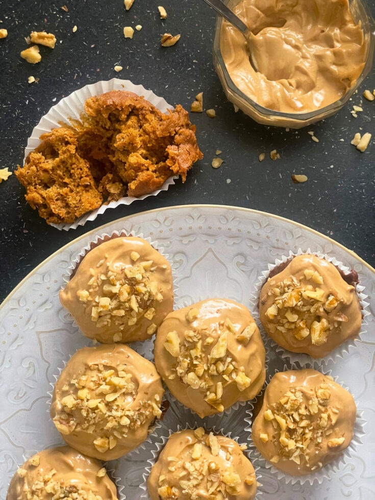 Torn carrot cake muffin and iced carrot cake muffins on decorative plate and a bowl of coconut sugar cream cheese icing with a spoon and chopped walnuts on a black counter