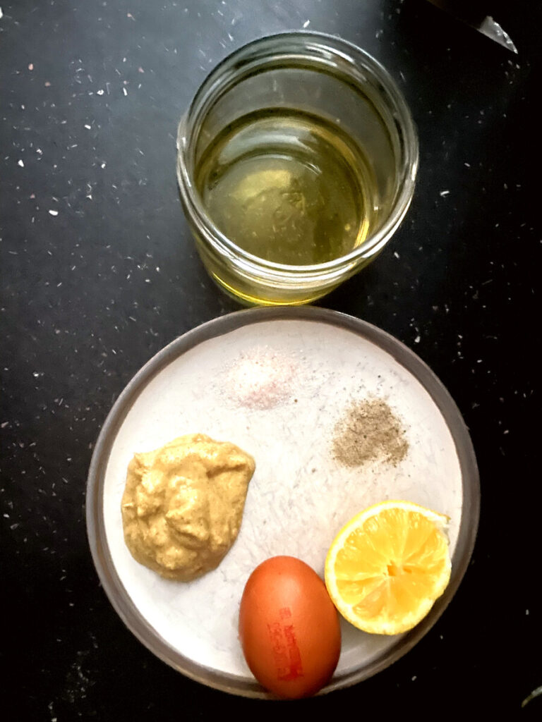 All ingredients for homemade avocado oil mayonnaise laid out: egg, mustard, lemon, salt, pepper and avocado oil